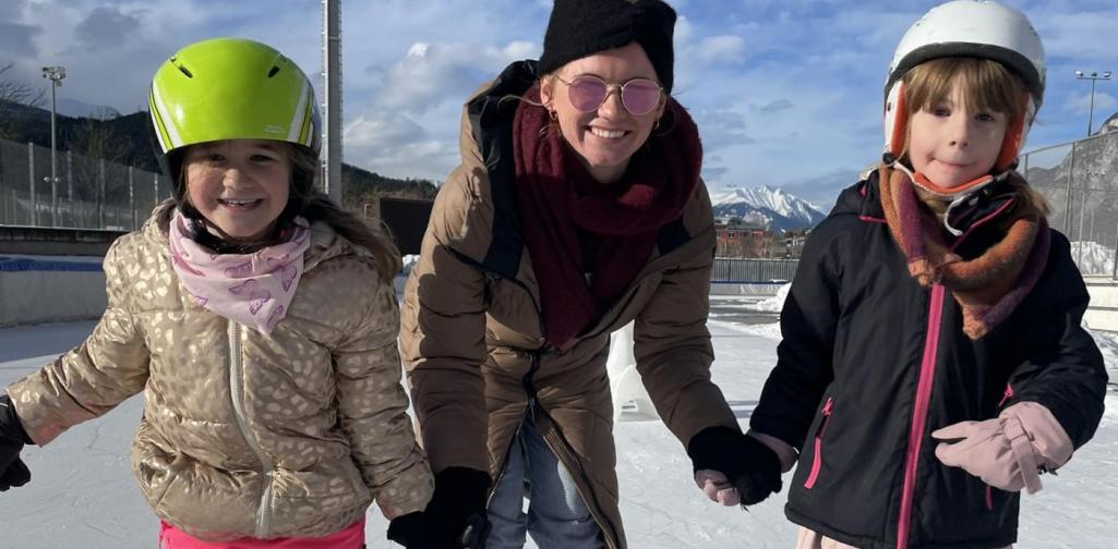 Begleitperson unterstützt zwei Kinder beim Schlittschuhlaufen auf einer Eisfläche im Freien.