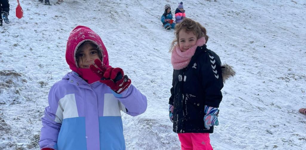Zwei Kinder stehen im Schnee am Amraser Hügel. Ein Kind hält ein rotes Rutschblatt und zeigt lächelnd ein Peace-Zeichen, das andere Kind steht daneben und lächelt in die Kamera. Im Hintergrund rutschen viele weitere Kinder den verschneiten Hügel hinunter.