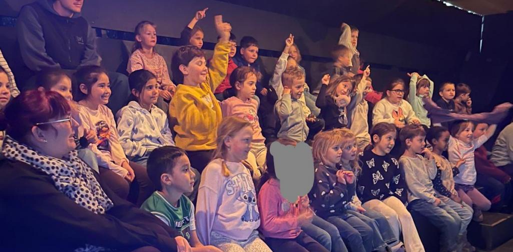 Stephanie Knabl Eine Gruppe von Grundschulkindern sitzt auf gestuften Sitzreihen in einem Theater- oder Veranstaltungsraum. Viele Kinder melden sich begeistert mit erhobenen Armen und schauen aufmerksam nach vorne. Eine erwachsene Begleitperson sitzt am Rand der Gruppe. Warmes Bühnenlicht beleuchtet die Szene und unterstreicht die lebendige, neugierige Atmosphäre.