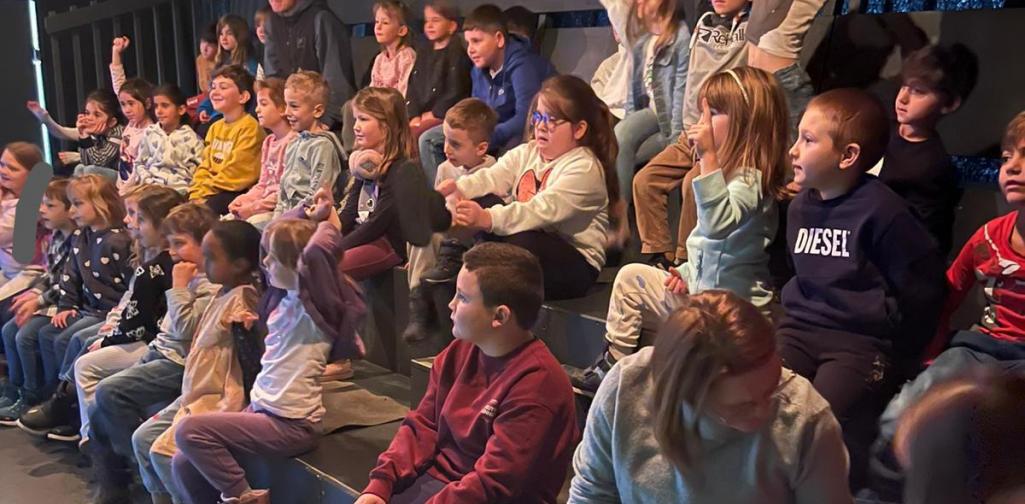 Stephanie Knabl Eine Gruppe von Grundschulkindern sitzt auf gestuften Sitzreihen in einem Theater- oder Veranstaltungsraum. Viele Kinder melden sich begeistert mit erhobenen Armen und schauen aufmerksam nach vorne. Eine erwachsene Begleitperson sitzt am Rand der Gruppe. Warmes Bühnenlicht beleuchtet die Szene und unterstreicht die lebendige, neugierige Atmosphäre.