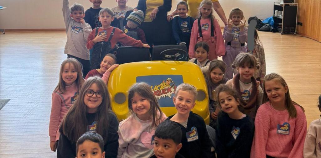 Eine große Gruppe von Kindern und zwei Erwachsene stehen und knien in einem hellen Raum um ein gelbes Lernfahrzeug „Blick und Klick“. Im Fahrzeug sitzt eine Stoffpuppe, die Kinder lächeln in die Kamera.