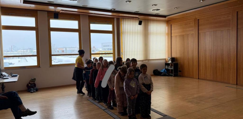 Mehrere Kinder stehen in einer Reihe an einem Übungs-Zebrastreifen in einem großen Raum. Eine Erwachsene begleitet die Gruppe während einer Verkehrssicherheitsübung.