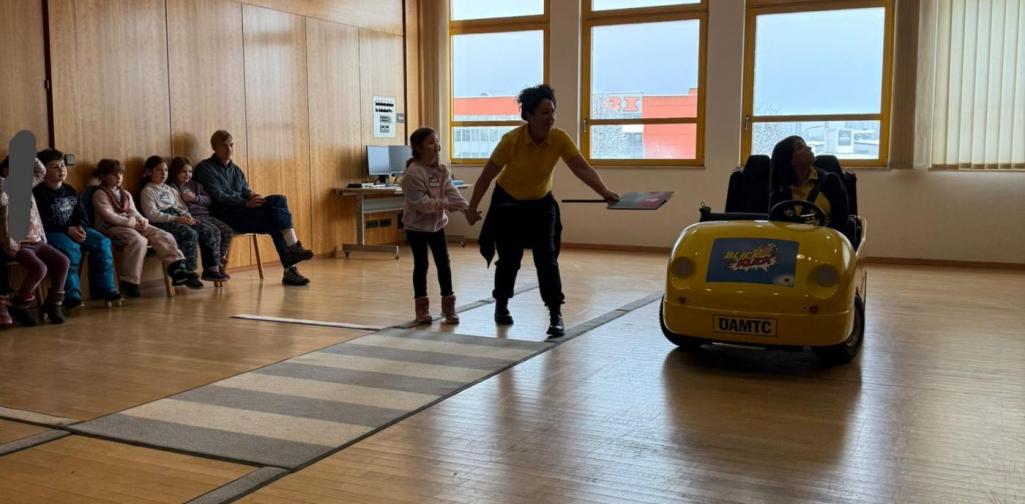 Eine Erwachsene begleitet ein Kind beim Überqueren eines Übungs-Zebrastreifens, während ein gelbes Lernfahrzeug „Blick und Klick“ anhält. Weitere Kinder sitzen im Hintergrund und beobachten die Szene.