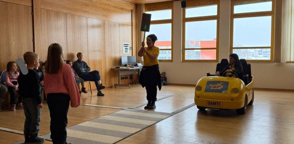 Eine Erwachsene hält ein grünes Fußgängersignal hoch, während Kinder am Übungs-Zebrastreifen warten. Daneben steht ein gelbes Lernfahrzeug zur Verkehrserziehung.