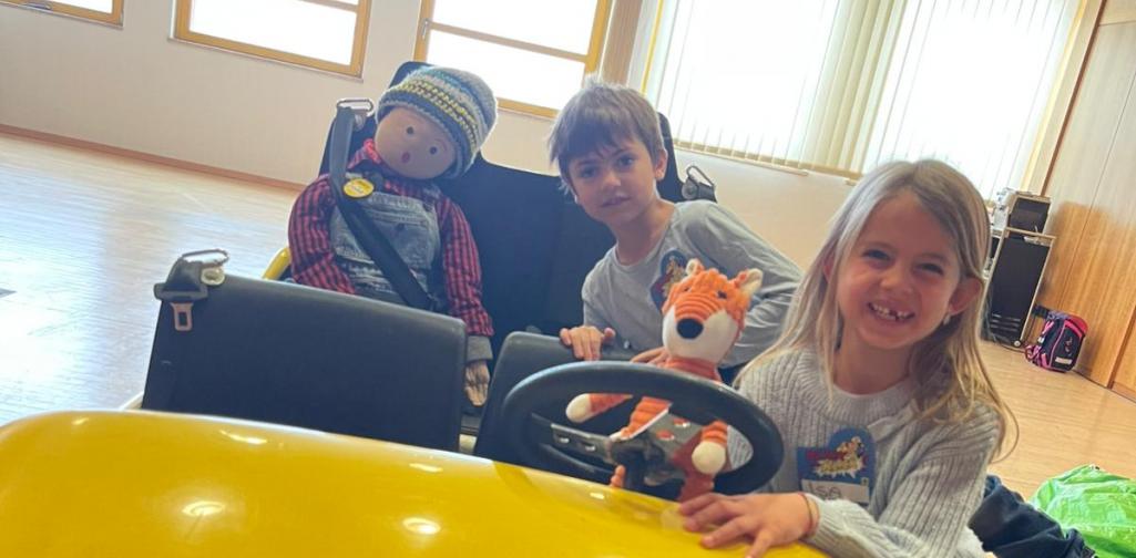 Drei Kinder sitzen und stehen in einem gelben Spielauto mit der Aufschrift „Blick und Klick“. Ein Stofffuchs sitzt am Lenkrad, während die Kinder lächelnd spielerisch richtiges Verhalten im Straßenverkehr üben.