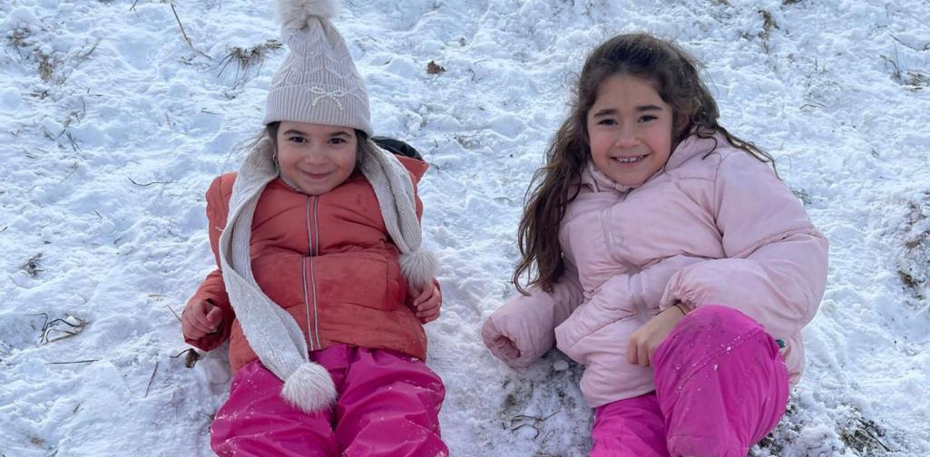Zwei Kinder liegen entspannt im Schnee auf dem Hang und lächeln fröhlich.
