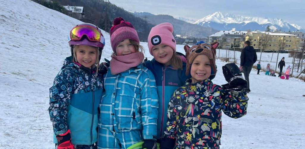 Vier Kinder stehen nebeneinander im Schnee und halten bunte Rutschblätter. Im Hintergrund sieht man Berge und weitere spielende Kinder.