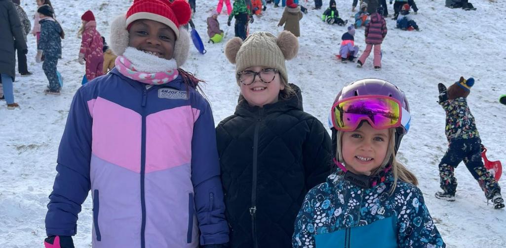 Drei Kinder stehen lächelnd vor dem verschneiten Hügel. Hinter ihnen rutschen viele Kinder mit Rutschblättern den Hang hinunter.