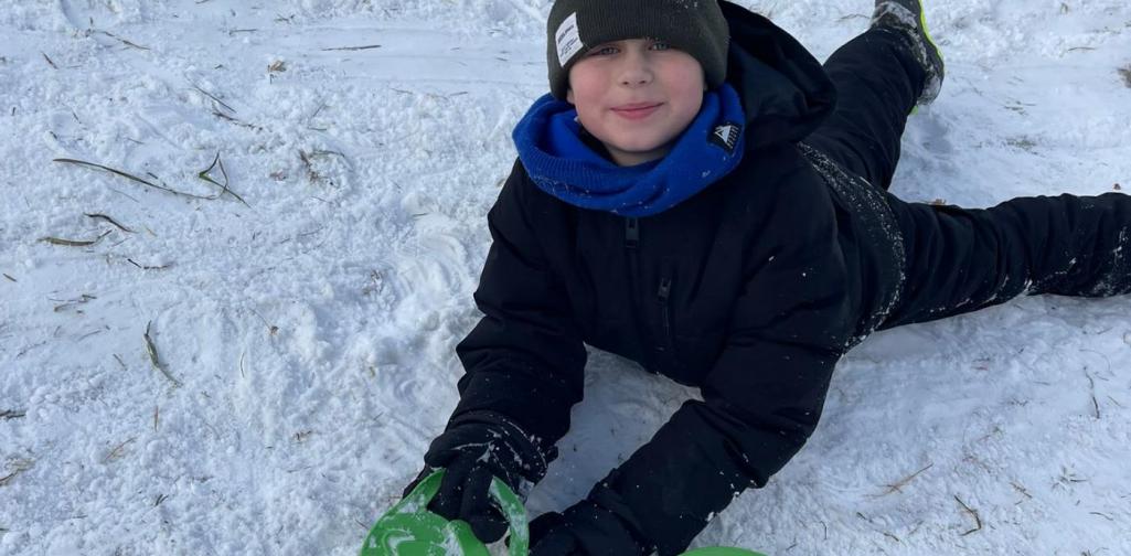 Ein Kind liegt bäuchlings im Schnee und hält ein grünes Rutschblatt vor sich.