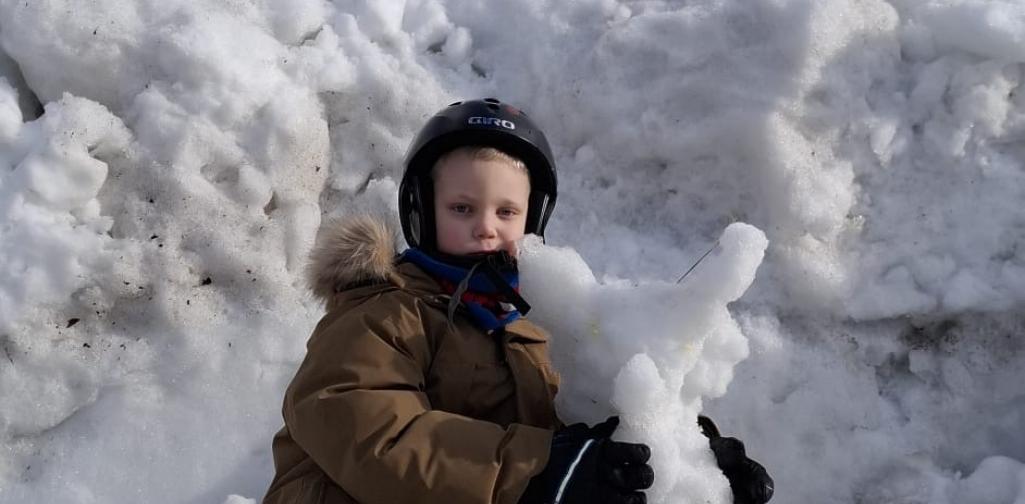 Ein Kind in Winterkleidung sitzt vor einem großen Schneehaufen und hält eine selbstgebaute Schneefigur im Arm. Das Kind trägt Helm und Handschuhe, der Himmel ist blau.
