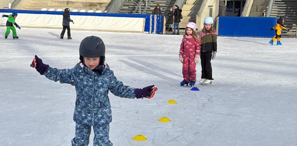 Ein Kind mit Helm fährt auf Schlittschuhen über eine Eisfläche und balanciert mit ausgebreiteten Armen durch einen Parcours aus Markierungshütchen.
