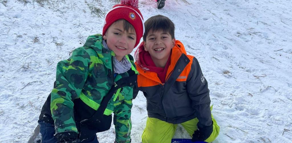 Zwei Kinder knien im Schnee neben einem blauen Rutschblatt und lachen in die Kamera.