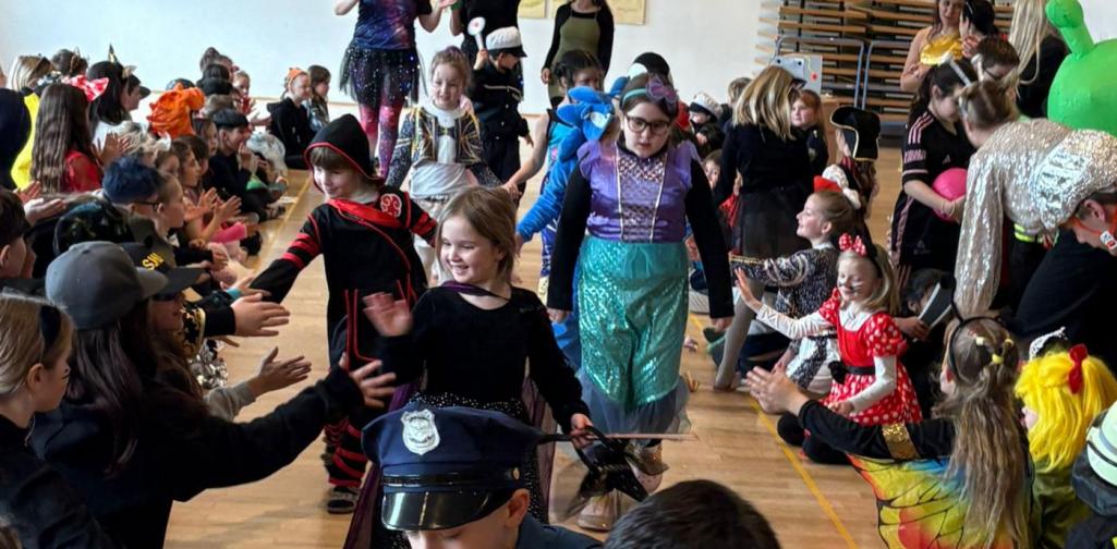 Kinder in bunten Faschingskostümen laufen durch eine Turnhalle Spalier, während Mitschülerinnen und Mitschüler am Rand sitzen, klatschen und High-Fives geben; im Vordergrund sind verkleidete Kinder als Polizisten zu sehen.