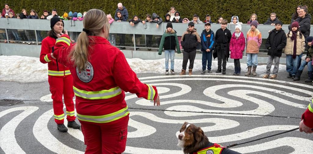 Zwei Rettungshundeführerinnen erklären den Schülerinnen und Schülern die Arbeit eines Rettungshundes, der in Einsatzkleidung an der Leine steht.