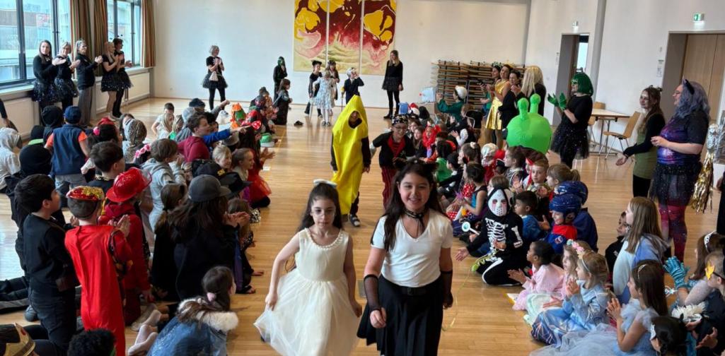 Zwei Mädchen gehen durch ein Spalier von verkleideten Mitschülerinnen und Mitschülern in einem Gemeindesaal, während Kinder und Lehrkräfte am Rand sitzen, klatschen und die Faschingskostüme bewundern.