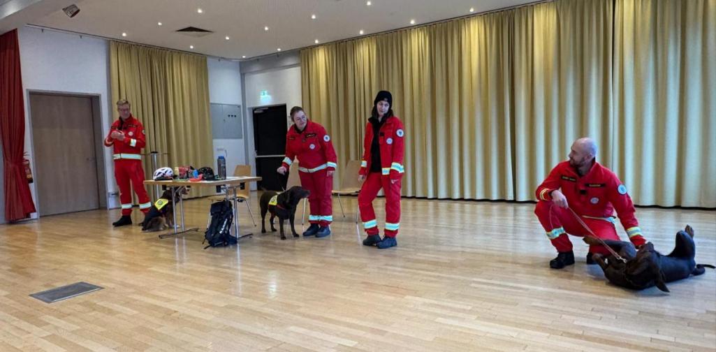 Mitglieder der Rettungshundestaffel zeigen in der Aula verschiedene Hunde und Ausrüstungsgegenstände und demonstrieren den Umgang mit den Tieren.