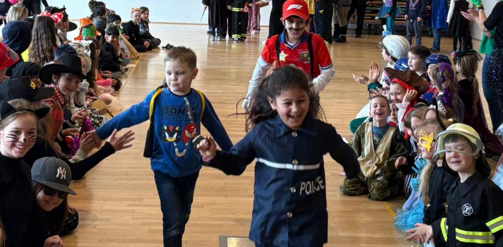 Drei verkleidete Kinder laufen fröhlich durch ein Spalier jubelnder Mitschülerinnen und Mitschüler in einem Gemeindesaal, während die sitzenden Kinder am Rand klatschen und High-Fives geben.
