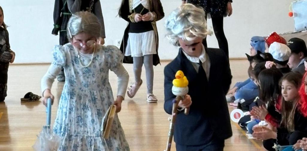 Zwei Kinder verkleidet als ältere Menschen gehen mit Gehstock und Regenschirm durch ein Spalier klatschender Mitschülerinnen und Mitschüler in einem Gemeindesaal.