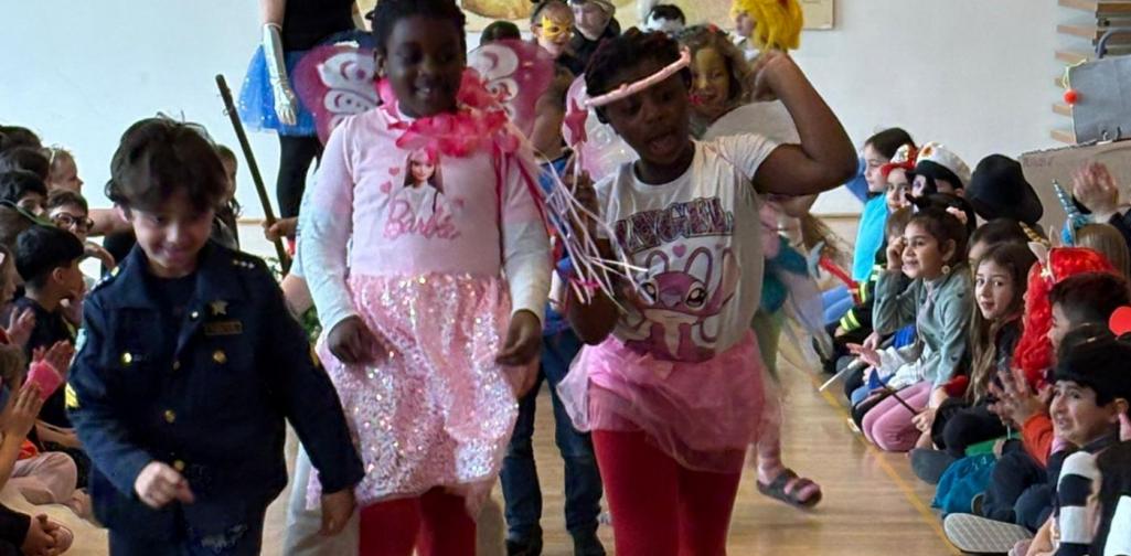 Drei Kinder in bunten Kostümen – darunter Fee und Prinzessin – laufen fröhlich durch ein Spalier klatschender Mitschülerinnen und Mitschüler in einem Gemeindesaal.