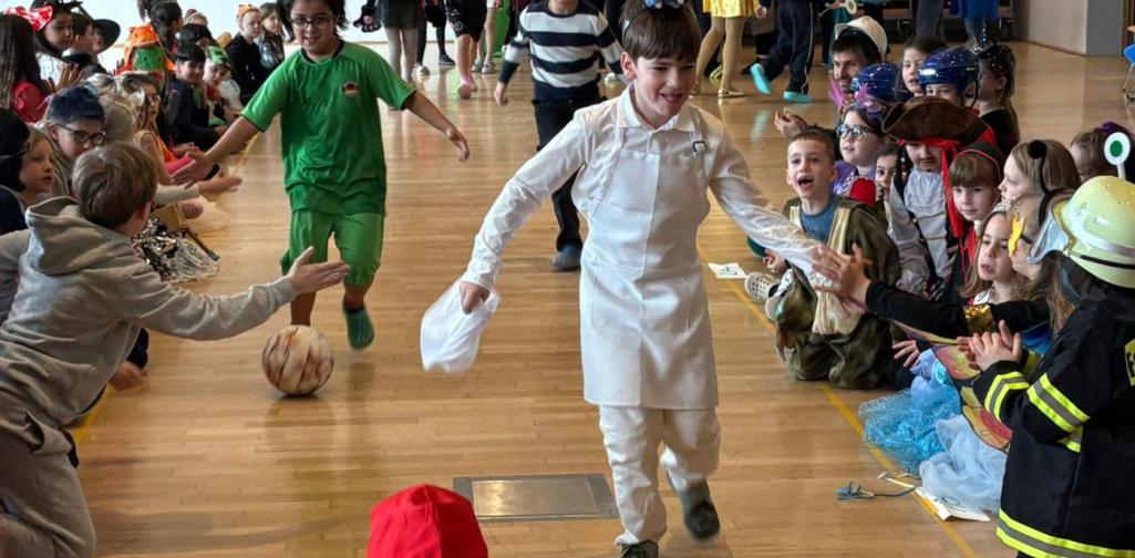Verkleidete Kinder laufen durch ein Spalier klatschender Mitschülerinnen und Mitschüler in einem Gemeindesaal; im Vordergrund kniet ein Kind im roten Ninja-Kostüm, während ein Junge in weißer Kleidung vorbeiläuft.