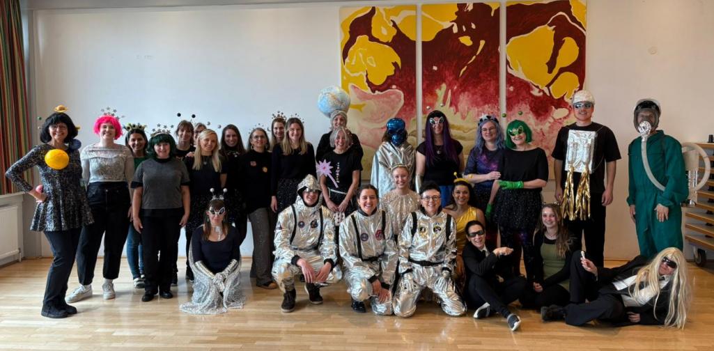 Das Lehrpersonal posiert verkleidet für ein Gruppenfoto in einem Gemeindesaal; die Kostüme reichen von Astronauten und Außerirdischen bis zu fantasievollen und glitzernden Verkleidungen.