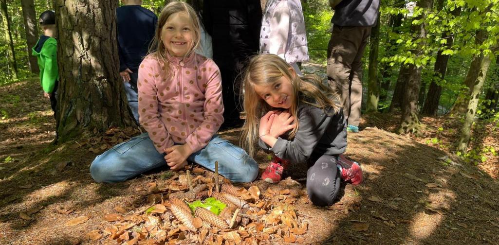„Zwei Schülerinnen knien im Wald neben einem aus Tannenzapfen und Blättern gestalteten Muster auf dem Boden.“