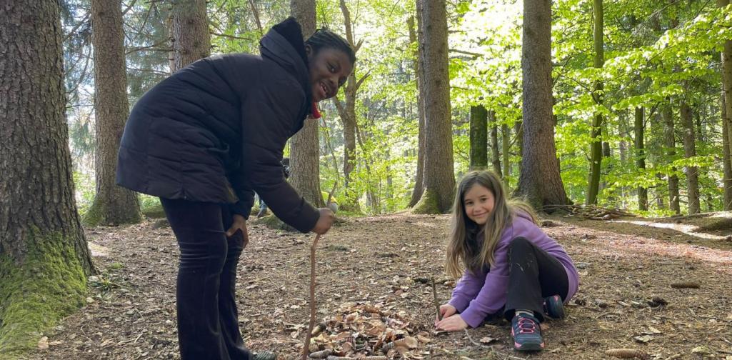 „Zwei Schülerinnen gestalten im Wald mit Stöcken, Blättern und Tannenzapfen ein Muster auf dem Waldboden.“