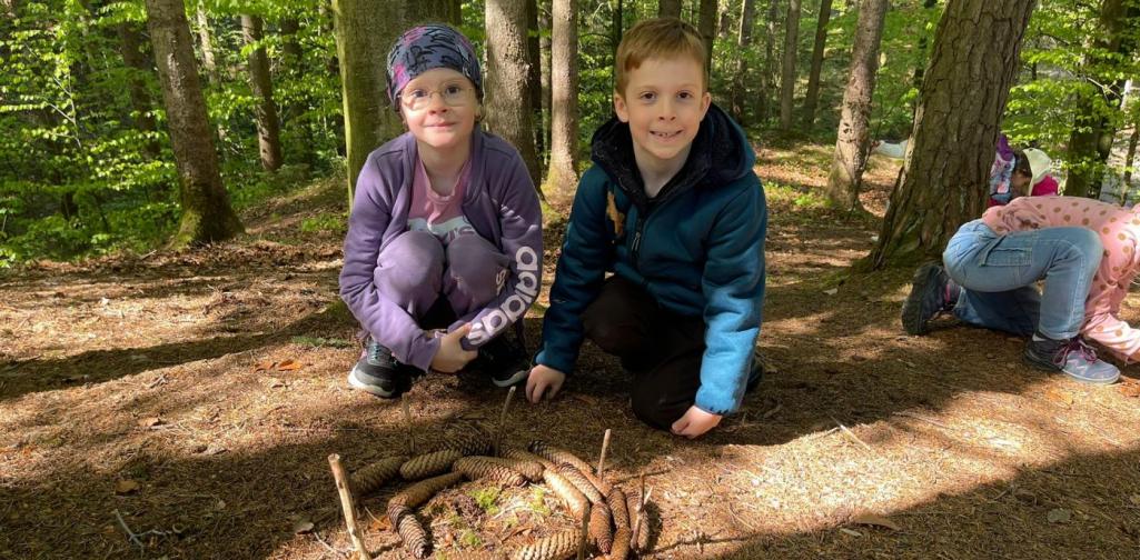 „Zwei Schüler knien im Wald neben einem Kreis aus Tannenzapfen und kleinen Stöcken, den sie gestaltet haben.“