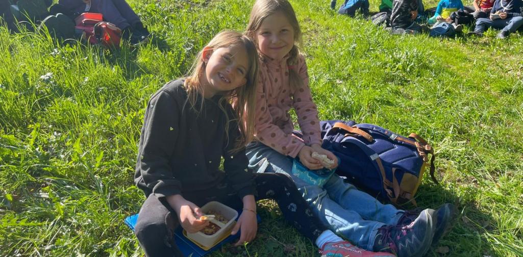 „Zwei Schülerinnen sitzen auf einer Wiese und essen während einer Pause im Freien, weitere Kinder sitzen im Hintergrund.“