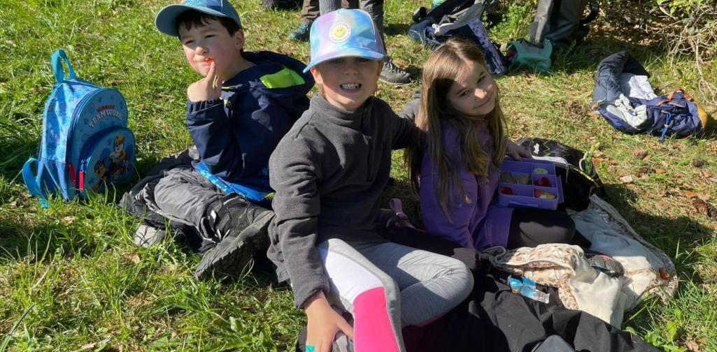 „Drei Schüler sitzen auf einer Wiese und essen gemeinsam ihre Jause während eines Ausflugs.“