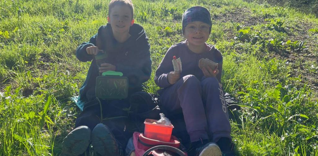 „Zwei Schüler sitzen auf einer Wiese und essen ihre Jause während eines Ausflugs im Freien.“