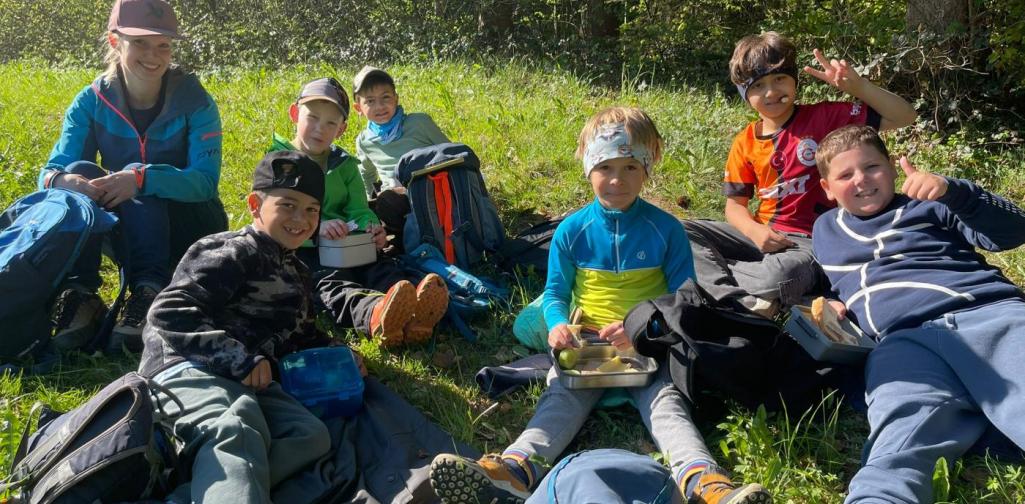 „Schüler:innen sitzen gemeinsam auf einer Wiese und essen ihre Jause während eines Ausflugs im Freien.“
