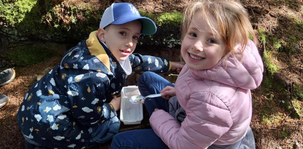 „Zwei Kinder sitzen im Wald auf dem Boden und untersuchen mit Becher und Löffel eine kleine Probe. Neben ihnen liegen Becherlupen und Materialien, während sie neugierig in die Kamera schauen und lächeln.“
