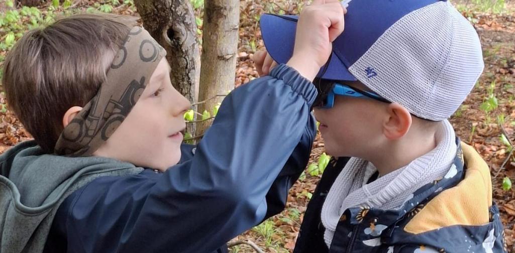 „Zwei Kinder stehen im Wald einander gegenüber, während eines dem anderen vorsichtig etwas auf die Kappe setzt oder zeigt. Im Hintergrund sind Bäume und Laub zu sehen, die Szene wirkt ruhig und konzentriert.“