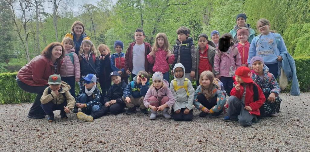 „Eine Schulklasse mit zwei Erwachsenen posiert gemeinsam für ein Gruppenfoto im Grünen. Die Kinder stehen und hocken auf einem Weg, umgeben von Bäumen und frischem Laub.“