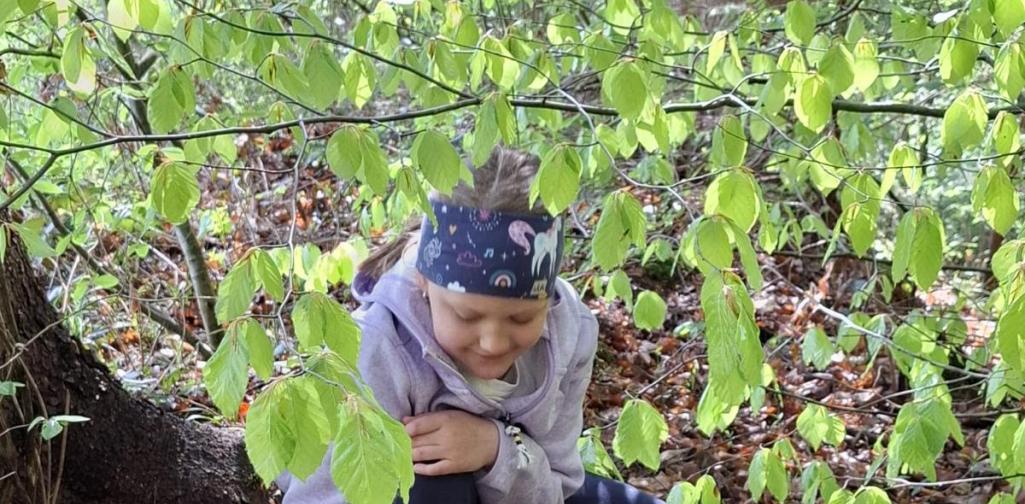 „Zwei Kinder knien im Wald unter grünen Zweigen und sammeln Naturmaterialien vom Boden. Ein Kind hält eine kleine Schale bereit, während beide konzentriert den Waldboden untersuchen.“