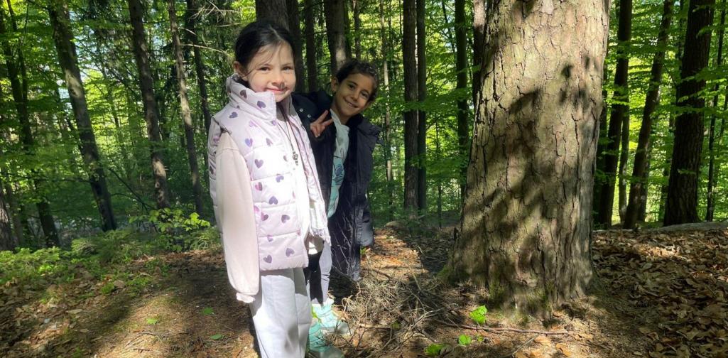 „Zwei Schülerinnen stehen im Wald neben einem Baum und zeigen gesammelte Äste und Naturmaterialien am Boden.“