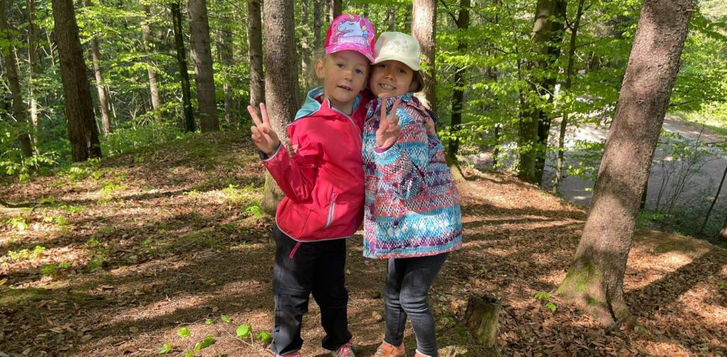 „Zwei Schülerinnen stehen im Wald neben einem Kreis aus Tannenzapfen und Blättern und zeigen mit den Händen ein Friedenszeichen.“