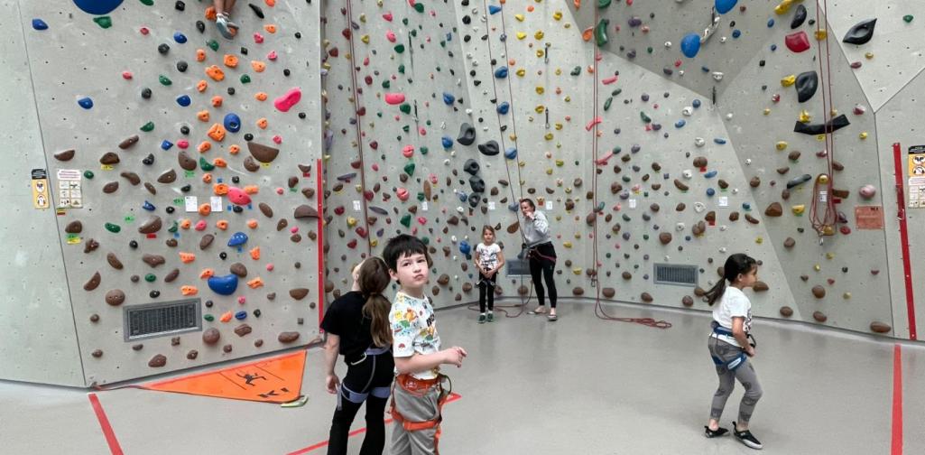 Kinder mit Klettergurten bewegen sich in einer Kletterhalle, während ein Kind eine Kletterroute an der hohen Kletterwand erklimmt.