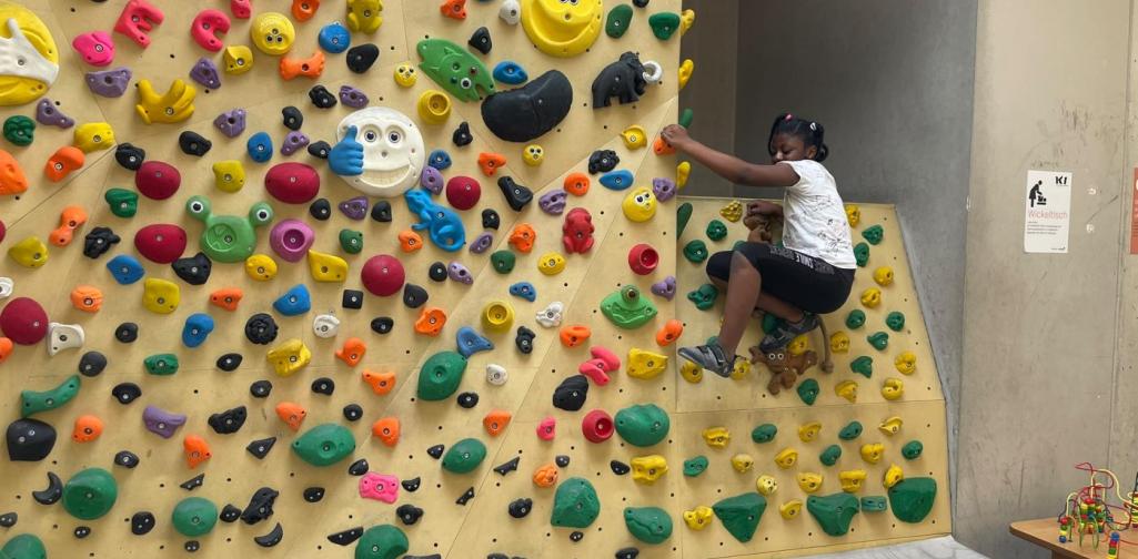 Ein Kind klettert an einer bunten Boulderwand und probiert verschiedene Griff- und Trittkombinationen aus.