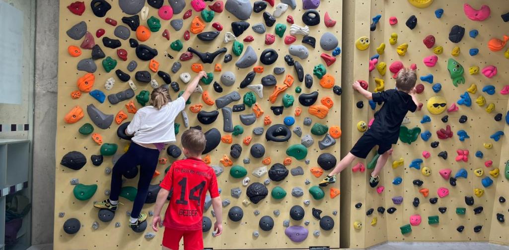 Kinder bouldern an einer Indoor-Kletterwand mit unterschiedlich geformten und farbigen Griffen.