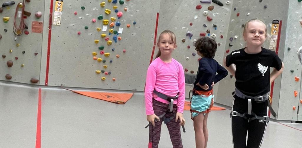 Kinder mit Kletterausrüstung stehen in einer Kletterhalle vor einer Kletterwand, während ein Kind eine Route erklimmt.