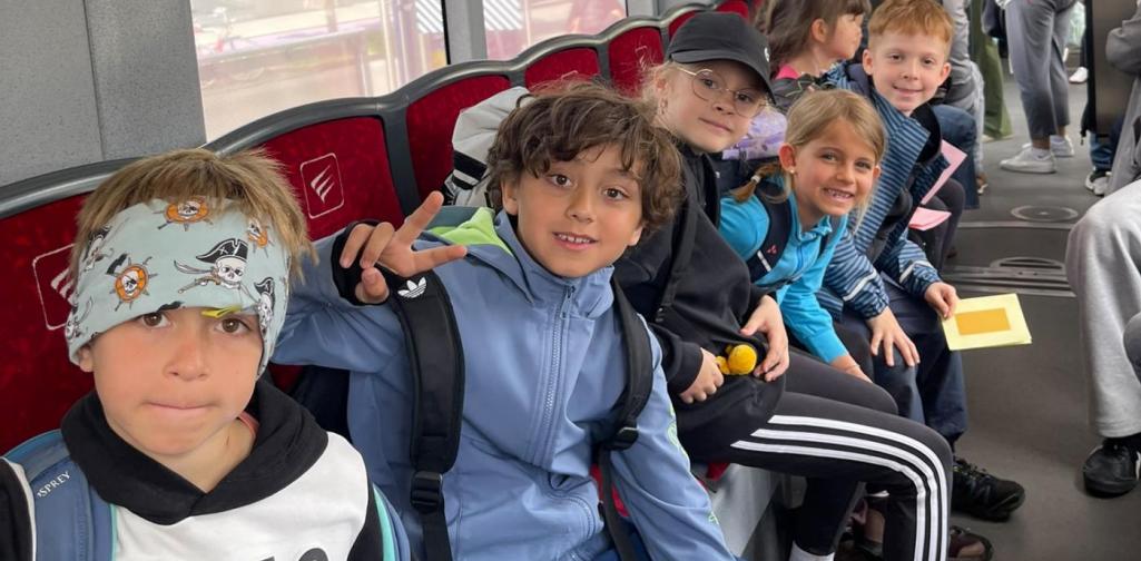 VS Amras Kinder einer Schulklasse sitzen während einer Exkursion in einer Straßenbahn und halten Arbeitsmaterialien für eine Lernaufgabe bereit.