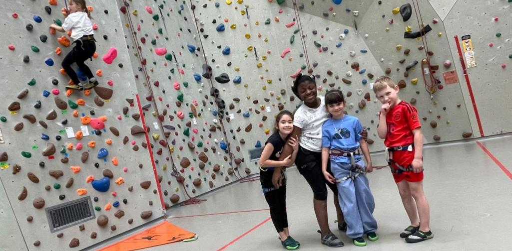 Kinder mit Klettergurten stehen in einer Kletterhalle vor einer Kletterwand, während ein Kind eine Route an der Wand erklimmt.
