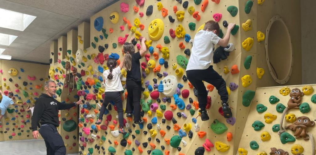 Kinder bouldern an einer farbenfrohen Kletterwand, während ein Trainer sie unterstützt und begleitet.