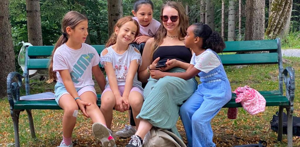 Eine Lehrperson sitzt mit vier Kindern auf einer Parkbank und unterhält sich mit ihnen im Schlosspark.
