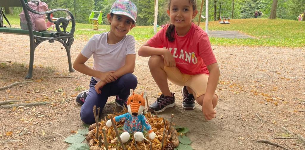 Zwei Kinder präsentieren eine Naturkunst aus Blättern, Zapfen und einem Stofftier auf dem Boden des Schlossparks.