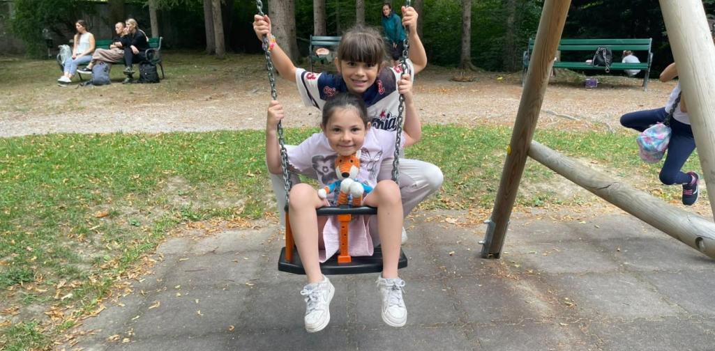 Zwei Kinder schaukeln gemeinsam auf einer Schaukel und haben sichtlich Freude am Spielen im Schlosspark.“