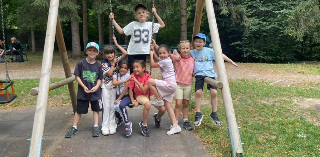 Eine Gruppe von Kindern versammelt sich lachend rund um eine Schaukel auf dem Spielplatz im Schlosspark.“