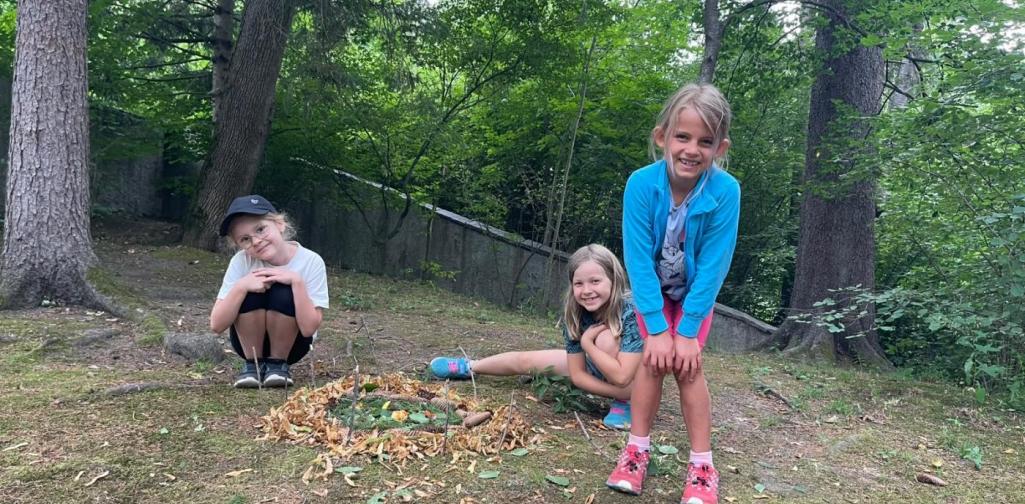 Drei Kinder präsentieren lächelnd ein selbst gestaltetes Naturkunstwerk aus Blättern, Zweigen und Naturmaterialien im Schlosspark.